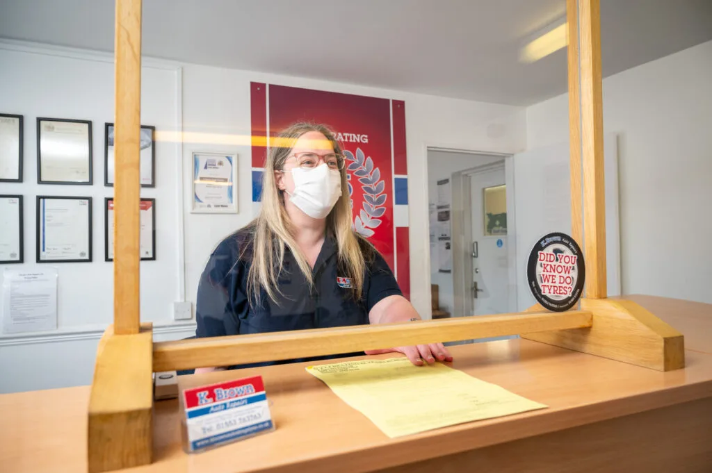 Receptionist wearing mask smiling behind desk and covid screen handing customer invoice